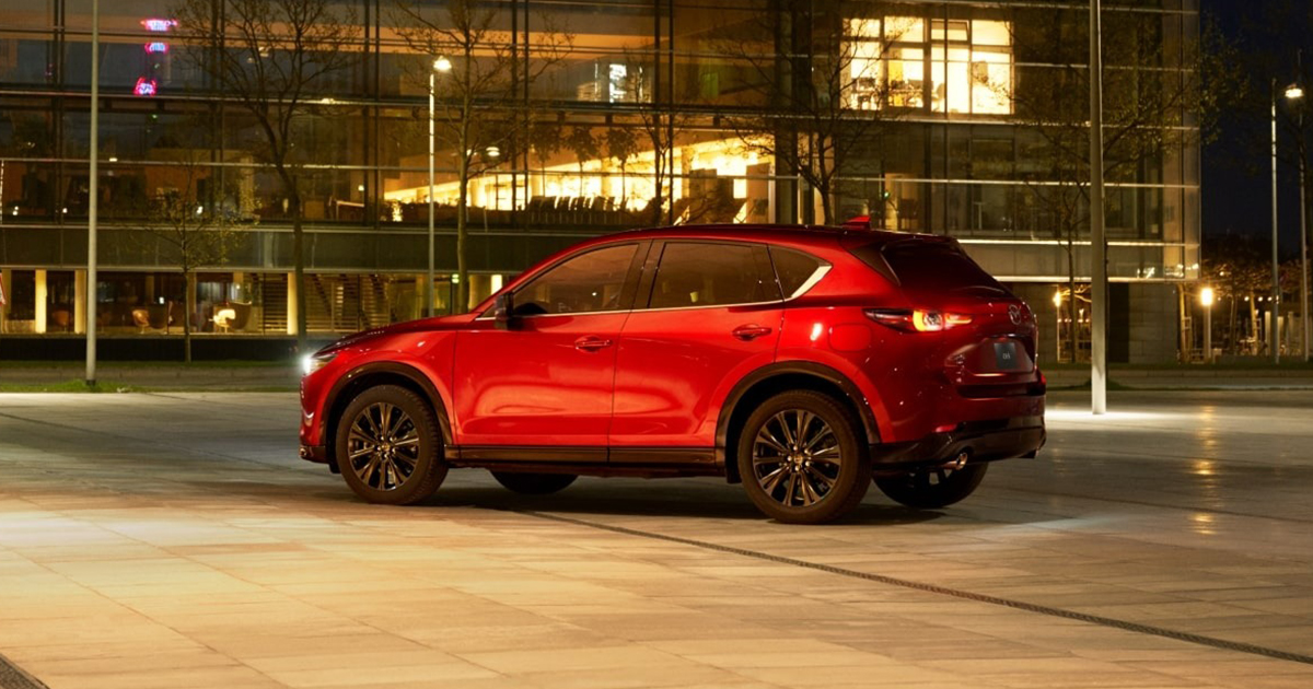 Red SUV parked in front of a glass building with lights shining inside.