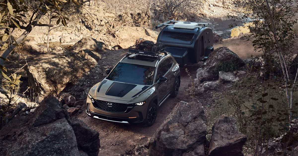 Mazda CX-50 SUV towing a compact camper trailer on a rugged dirt trail surrounded by large rocks and trees.