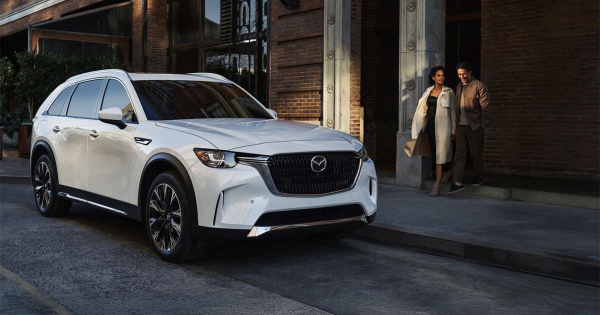 White Mazda CX-90 parked on city street with couple walking beside