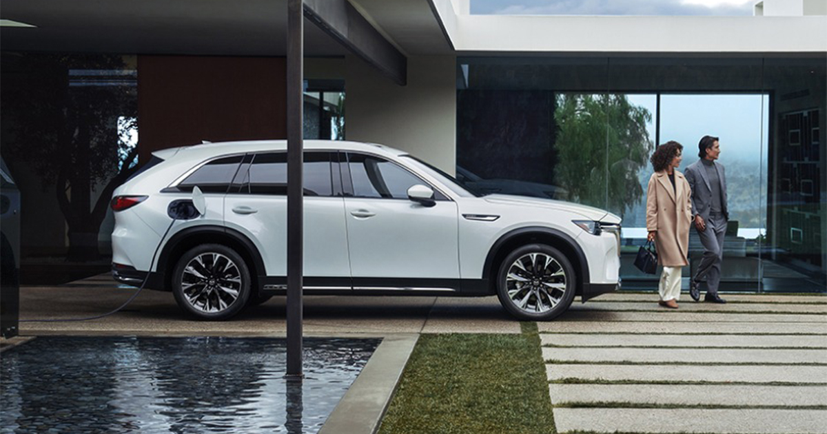 Side view of a white Mazda CX-90 PHEV charging at a home, with a modern glass-walled house and couple walking nearby.