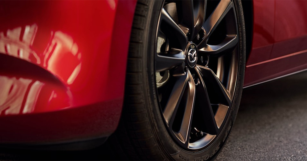 Close-up of a Mazda wheel with a black alloy rim and tire, with reflections of the car’s red body.
