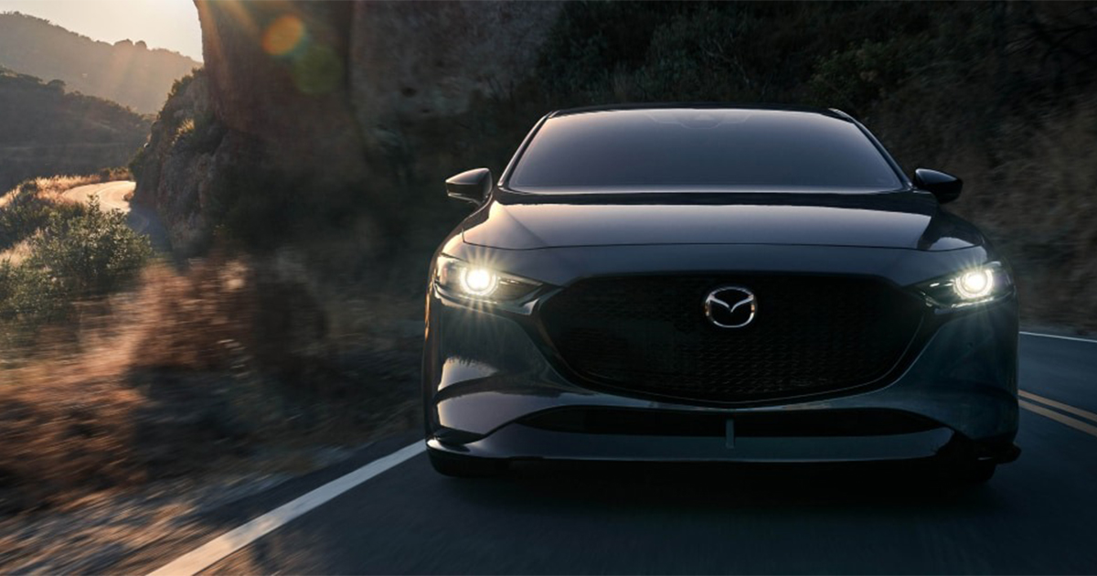 Front view of a black Mazda3 driving on a curvy road, headlights on and sun glare in the background.