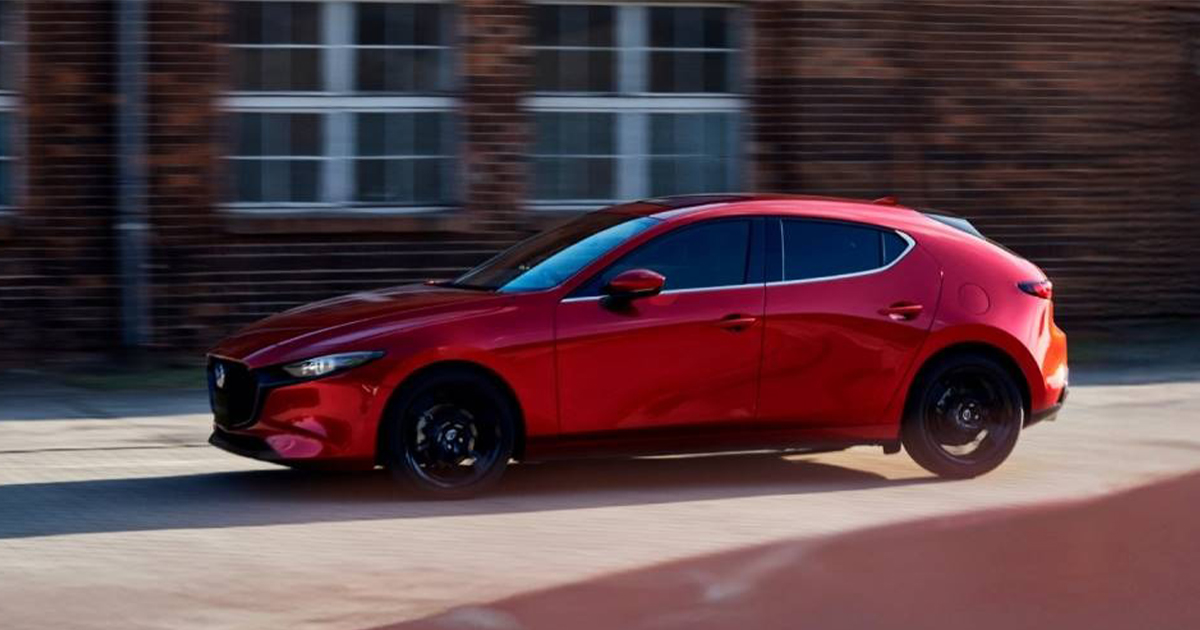 Side profile of a red Mazda3 hatchback in motion near a brick building, sunlight reflecting off the body.