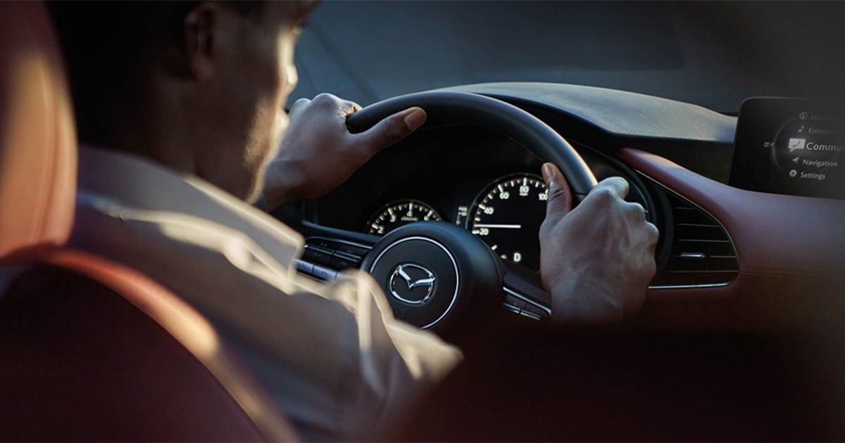Close-up of a driver holding the steering wheel of a Mazda, dashboard gauges and infotainment screen visible.