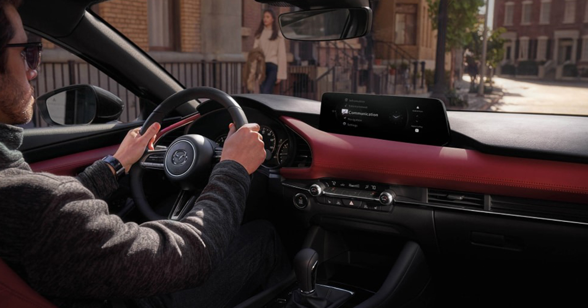 Driver’s view inside a Mazda interior with a red dashboard, showing the infotainment screen and steering wheel with Mazda logo.