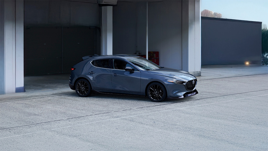 Side view of a gray Mazda3 hatchback parked outside a modern building with shutter doors.