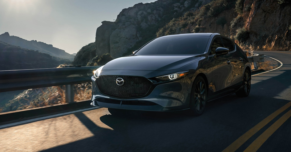 A black Mazda3 driving along a winding mountain road at sunset, headlights on and casting reflections.