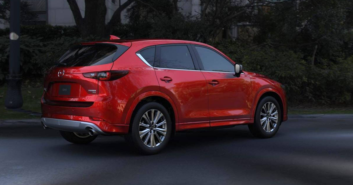 Rear three-quarter view of a red Mazda CX-5 parked on a shaded street, with trees and bushes in the background.
