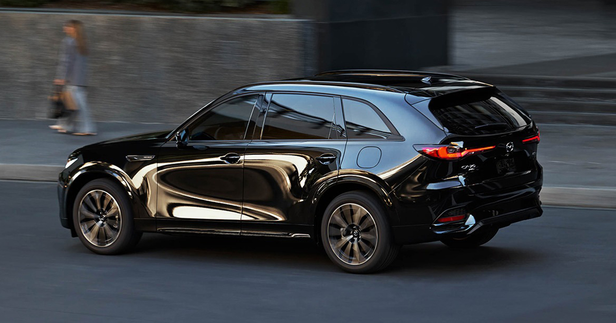 Side view of a black Mazda CX-90 SUV driving on a city street with reflections across its body.