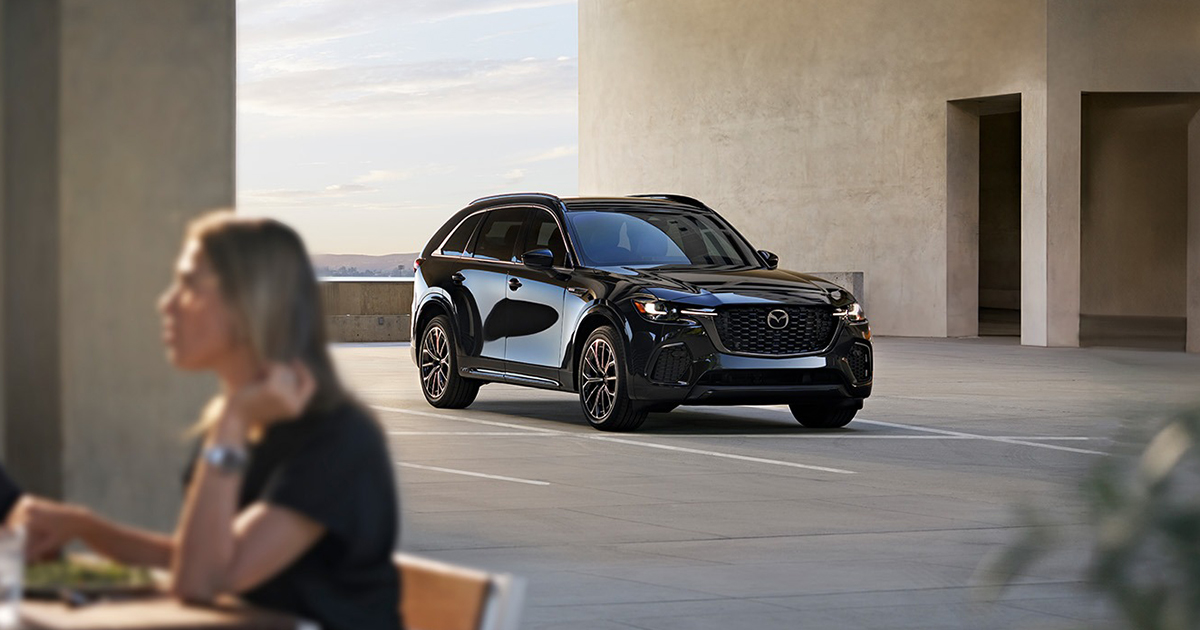 Black Mazda CX-90 SUV parked in a sunlit, modern concrete setting with a woman sitting blurred in the foreground.