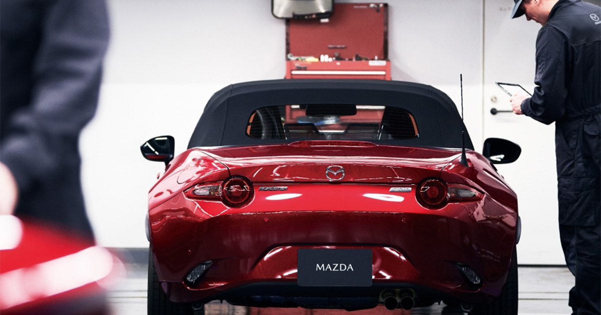 Red Mazda MX-5 Miata convertible parked inside a service bay with a technician inspecting the car.